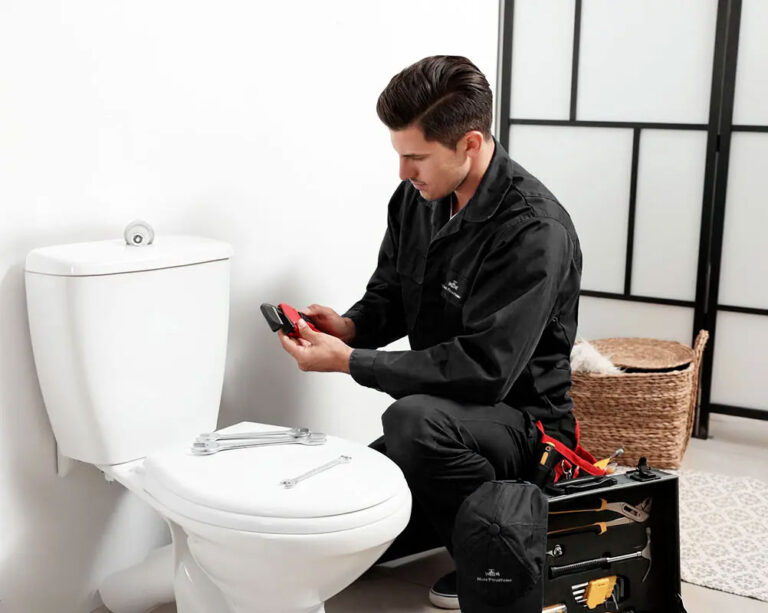 Plombier en noir assis devant des toilettes, tenant un outil pour réparation.